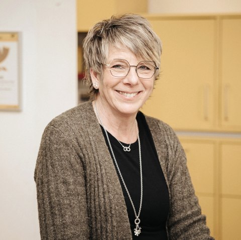 Eine Frau steht hinter einem Tresen eines Postschalters. Hinter ihr Schränke und das Posthorn-Logo.