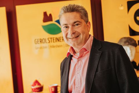 Ein schick gekleideter Herr mit Hemd und Sakko lächelt in die Kamera. Hinter ihm das Logo der Touristik GmbH Gerolsteiner Land und einige Souvenierartikel.
