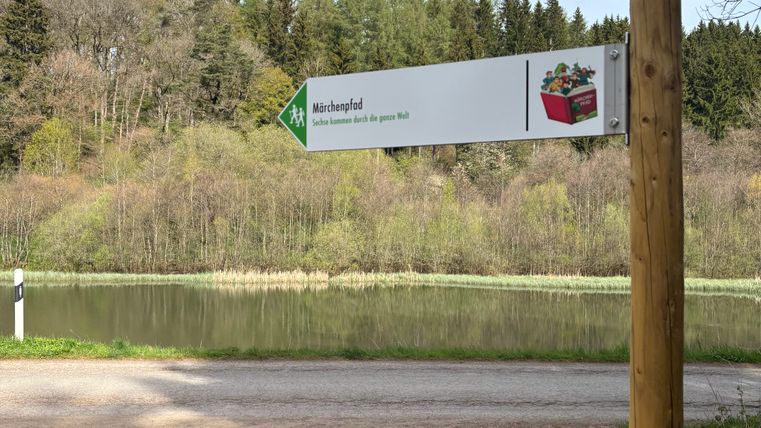 Ein Wegweiser im Wald zeigt den Märchenpfad an, mit einem See im Hintergrund.