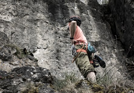 A person wearing a helmet and equipment stands in front of a steep rock face, hands on hips, looking up.