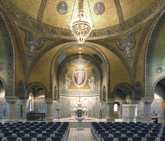 Das Innere der Erlöserkirche mit Blick zur Decke und zum Altar. Die Decke ist mit goldenen Mosaiken, Engeln und einem großen Kronleuchter geschmückt. Rundfenster lassen Licht in den prunkvollen Kirchenraum.