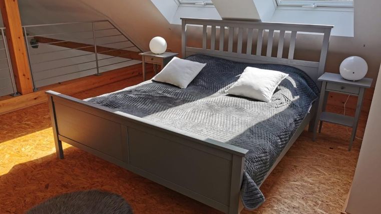 Een gezellige slaapkamer met een grijs bed en twee kussens. Grote ramen laten veel licht binnen en de vloer is van hout.