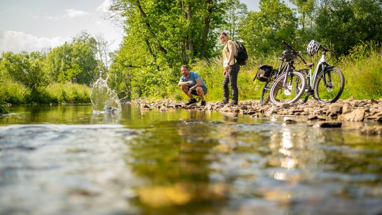 Zwei Personen stehen mit ihren Fahrrädern am Ufer eines Flusses inmitten einer grünen Parklandschaft