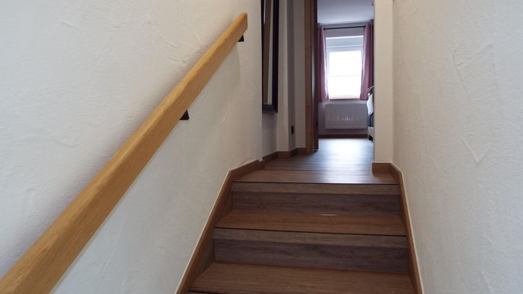 Ein heller Flur mit Holztreppe und einem Handlauf. Am Ende des Flurs ist ein Fenster mit Vorhängen zu sehen.