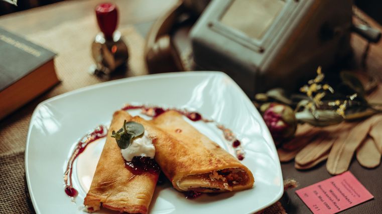 Ein Teller mit zwei gefüllten Crepes, garniert mit Sahne und einer fruchtigen Soße. Im Hintergrund sind dekorative Gegenstände und ein paar Bücher zu sehen.