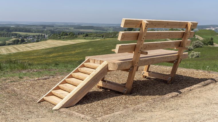 Eine übergroße Holzbank mit Treppe steht auf einem Hügel mit Blick auf Felder und Wälder unter blauem Himmel.