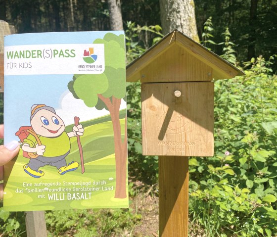 Das Prospekt "Wander(S)pass f&uuml;r Kids" wird hochgehalten vor einer dazugeh&ouml;rigen Stempelstation.
