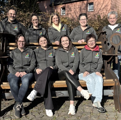 Een groep van 9 personen, waarvan sommigen op een bank zitten, lachen naar de camera. Alle personen dragen dezelfde jas met het bedrijfslogo erop geborduurd.