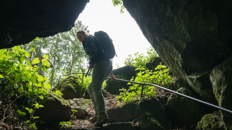 A woman at the exit of a dark cave climbs up the steps into the light and looks back into the cave.
