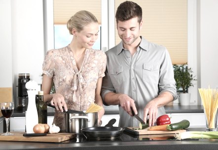 A couple stands side by side in the kitchen preparing a meal. Spaghetti is visible in the pot in front of them and vegetables are being chopped. 