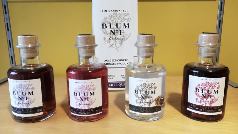 Verkaufsregal, auf dem vier Flaschen unterschiedlicher Sorten Gin der Marke "Blum N°1" stehen. Dahinter einige Flyer zu der Marke.