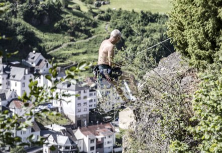 A shirtless man climbs a rock covered with plants, secured with ropes. Houses and green countryside can be seen in the background.