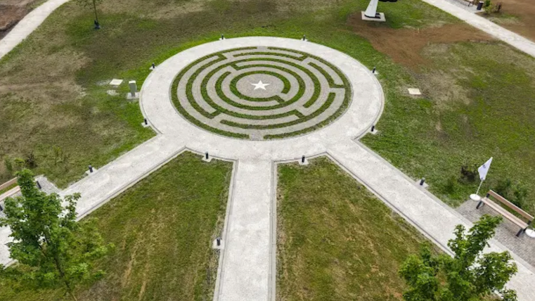 Luchtfoto van een landschapspark met een ronden labyrinth in het midden, omgeven door paden en gazons, evenals een op zonne-energie aangedreven bloemkunstwerk en zitgelegenheden.