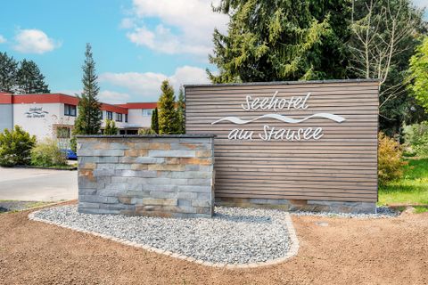 Ein Schild mit der Aufschrift „Seehotel au Strausee“ in einer grünen Umgebung. Im Hintergrund sind Bäume und ein Gebäude zu sehen.