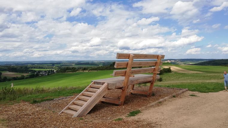 Eine riesige Holzbank mit Treppe steht in einer grünen Landschaft unter einem bewölkten Himmel. Ein Mensch ist am rechten Bildrand zu sehen.