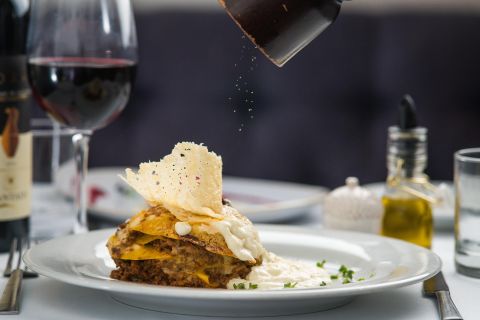 Lasagne auf einem Teller, garniert mit Käse und Kräutern. Im Hintergrund ein Glas Rotwein und eine Flasche Olivenöl. Gewürze werden darüber gestreut.