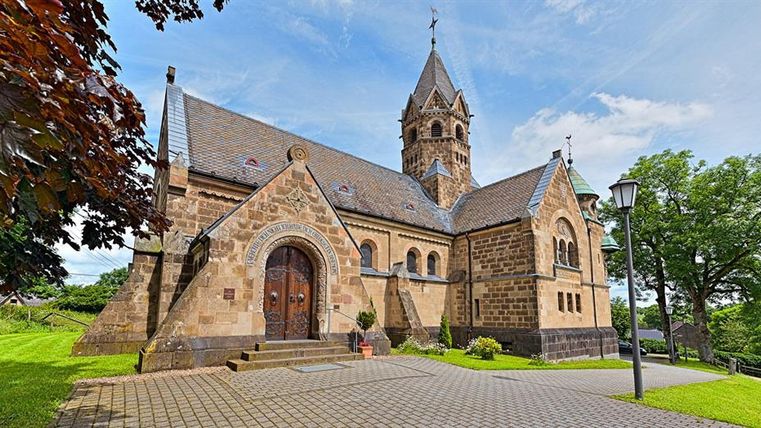 Die Erlöserkirche in Mirbach zeigt sich mit ihrem markanten Turm und Eingang. Umgeben von Bäumen und einem gepflasterten Weg, strahlt sie Ruhe aus.