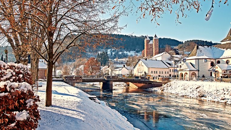 Ein verschneites Dorf, durch das ein Fluss fließt und eine Burg von oben thront.