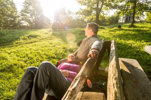 Un homme et une femme sur un banc dans une pelouse verte et luxuriante entourée d'arbres. La femme est allongée sur le dos, la tête sur les genoux de l'homme.