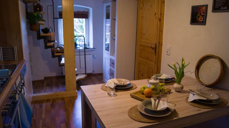 Een gezellige keuken met een houten tafel, gedekt met servies en fruit. Op de achtergrond zijn meubels en een trap zichtbaar die naar de volgende verdieping leidt.