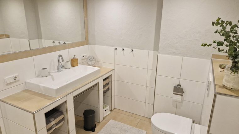 Ein modernes Badezimmer mit einem weißen Waschbecken und einem großen Spiegel. Die Wände sind hell gefliest und es gibt eine dezente Dekoration.