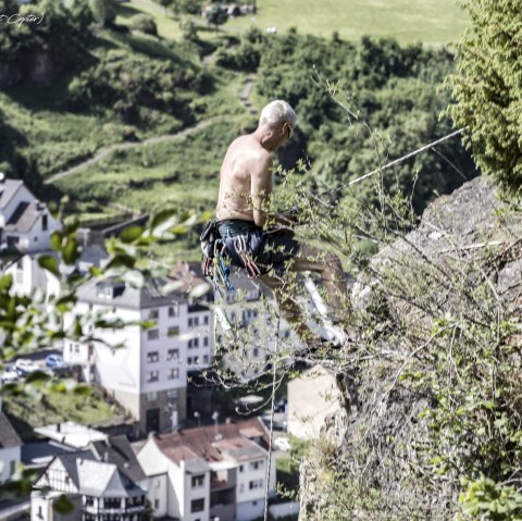 A shirtless man climbs a rock covered with plants, secured with ropes. Houses and green countryside can be seen in the background.