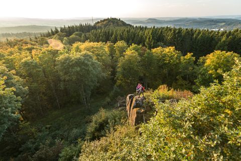 Zwei Personen stehen auf einem Felsen mit Blick auf einen Wald und Hügel im Hintergrund.