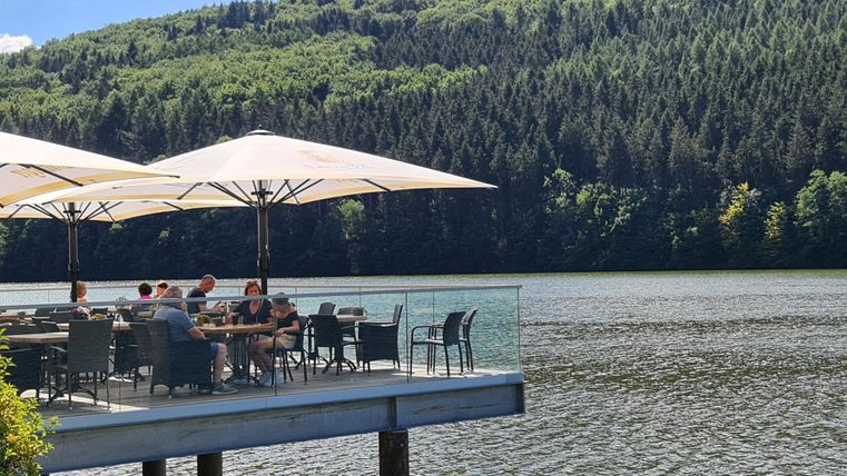 Un restaurant confortable au bord de l'eau avec des parasols et des clients appréciant la vue. En arrière-plan, on peut voir des collines boisées.
