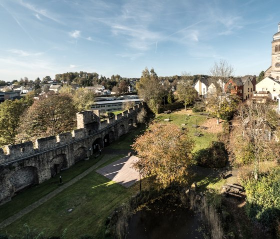 Panoramablick auf Hillesheim mit historischer Stadtmauer, grüner Parkanlage und einer Kirche im Hintergrund unter blauem Himmel., © Eifel Tourismus GmbH, D. Ketz