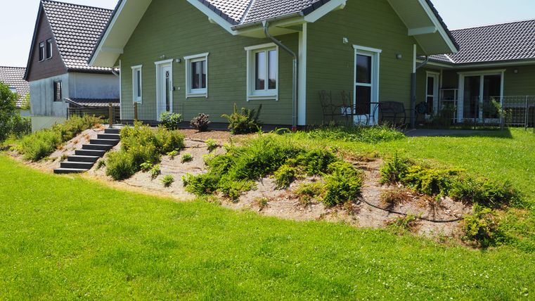 Ein modernes, grün gestrichenes Haus mit einem gepflegten Garten. Im Hintergrund sind weitere Wohnhäuser und ein blauer Himmel zu sehen.