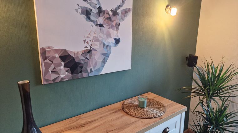 Ein modernes Wohnzimmer mit einem geometrischen Hirschbild an einer grünen Wand. Darunter steht ein Holztisch mit einer Blume, einer Kerze und einem eleganten schwarzen Vasen.