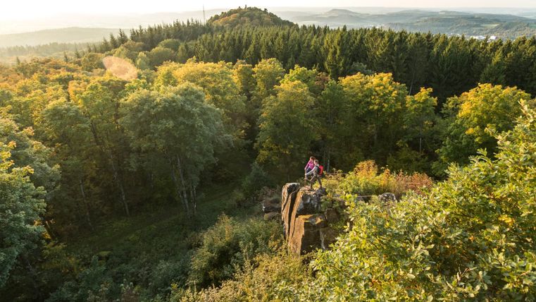 Zwei Personen stehen auf einem Felsen mit Blick auf einen Wald und Hügel im Hintergrund.
