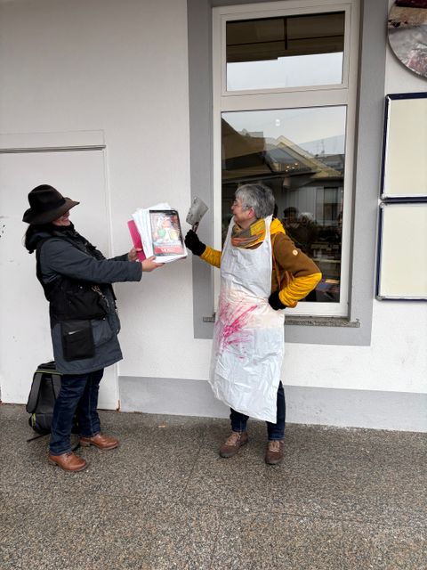 Eine dunkel gekleidete Person hält das Titelbild eines Eifelkrimis neben eine weiteren Person. Diese trägt eine blutige Metzgerschürze und hält ein Hackebeil hoch. 
