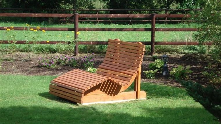 Une chaise longue en bois est posée sur une pelouse verte. En arrière-plan, une clôture en bois et quelques plantes sont visibles.