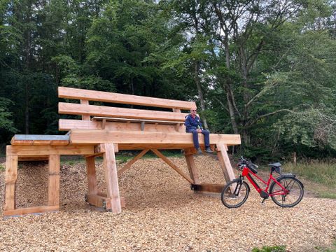 Eine Person sitzt auf einer übergroßen Holzbank im Freien neben einem roten Fahrrad.