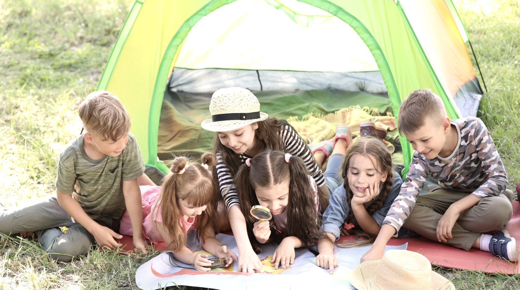 Sechs Kinder liegen und sitzen dicht beieinander vor einem bunten Zelt auf einer Wiese und schauen auf eine Landkarte, die vor ihnen liegt. Manche Kinder zeigen auf die Karte und ein Kind schaut genauer mit einer Lupe.