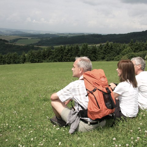 Drei Wanderer sitzen auf einer gr&uuml;nen Wiese und blicken in die Ferne. Einer tr&auml;gt einen orangefarbenen Rucksack. Im Hintergrund sind H&uuml;gel und W&auml;lder zu sehen., &copy; Eifel Tourismus GmbH
