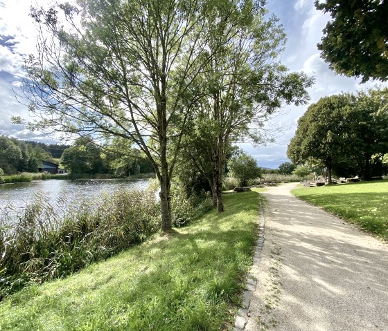 Ein sonniger Park mit einem geschwungenen Weg entlang eines Sees, umgeben von Bäumen und grünen Wiesen., © Touristik GmbH Gerolsteiner Land