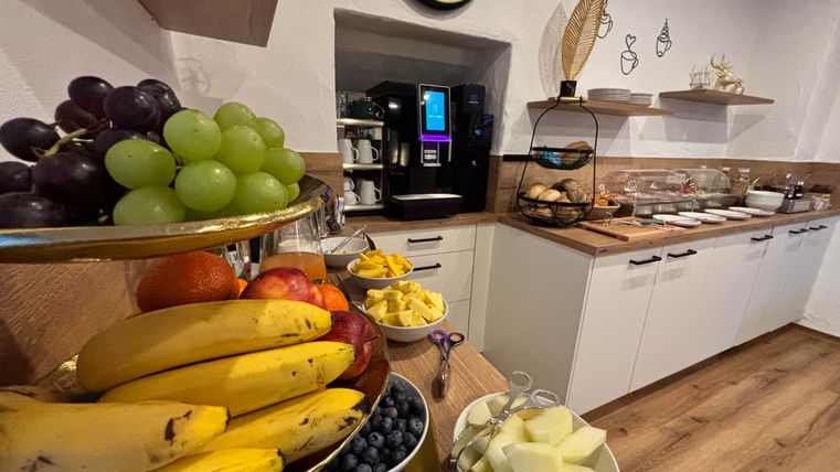 Eine gemütliche Küche mit einer Obstschale voller Bananen, Trauben und Äpfeln. Im Hintergrund steht ein Kaffeemaschine und eine Auswahl an Snacks auf Regalen.