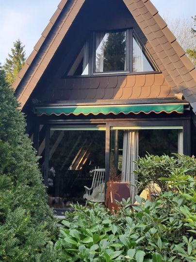 Ein gemütliches Holzhaus mit einem spitzen Dach. Das Haus ist von grünen Pflanzen umgeben und hat große Fenster.