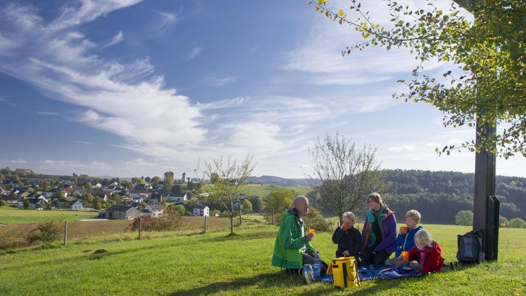 Familie beim Picknick auf einer Wiese mit Blick auf ein Dorf und blauen Himmel.