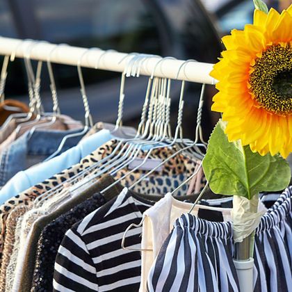 Eine Kleiderstange mit verschiedenen Kleidungsstücken, darunter gestreifte Tops und Hosen. Neben den Kleidern steht eine Sonnenblume, die das Bild auflockert.