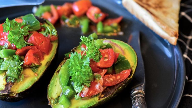 Gefüllte Avocadohälften mit Tomaten und Paprika auf einem Teller, daneben ein Stück Toast.