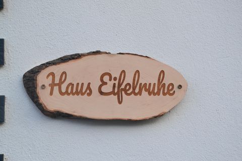 Ein Holzschild mit der Aufschrift „Haus Eifelruhe“. Es ist an einer weißen Wand befestigt.