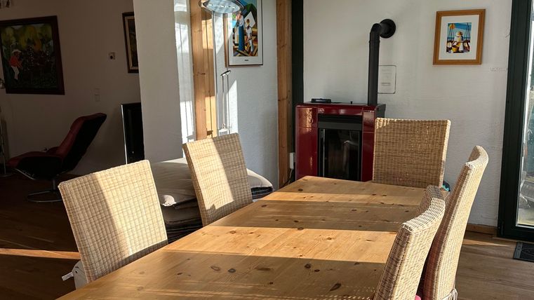 Ein helles Esszimmer mit einem großen Holztisch und mehreren Stühlen. Im Hintergrund sind gemütliche Möbel und ein Kamin zu sehen.
