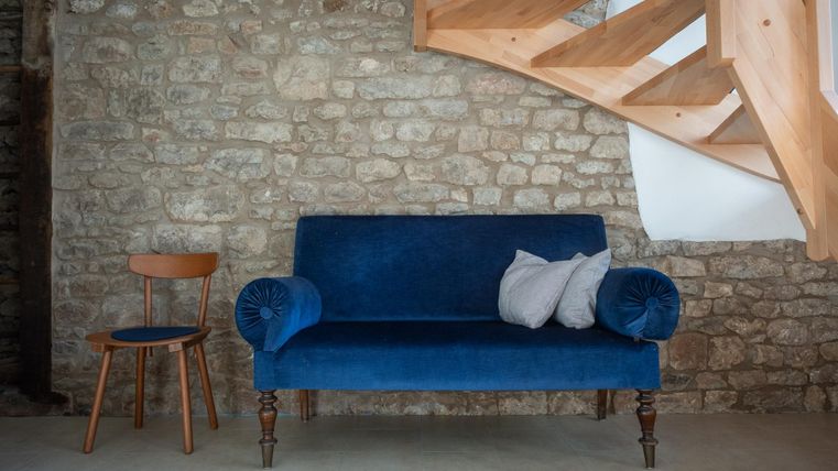 Ein stilvolles, blaues Sofa steht vor einer steinernen Wand. Neben dem Sofa befindet sich ein Holzstuhl und eine Treppe im Hintergrund.