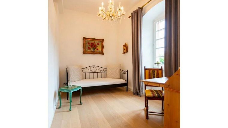 Ein einfaches Zimmer mit einem Bett, einem Tisch und Stühlen. Helle Wände und ein Kronleuchter sorgen für eine einladende Atmosphäre.