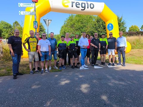 Zahlreiche Personen, welche teilsweise sportlicher Radfahrkleidung tragen, stehen auf dem Radweg unter einem großen Luftbogen des Sponsors e-regio.