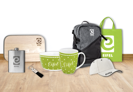 Een plankje, heupfles, sleutelhanger, schaal, mok, rugzak, stoffen tas en baseballpet naast elkaar op een houten vloer. Alle producten zijn afkomstig uit de Eifelshop en voorzien van het opschrift Eifel en/of het logo &ldquo;e&rdquo;.