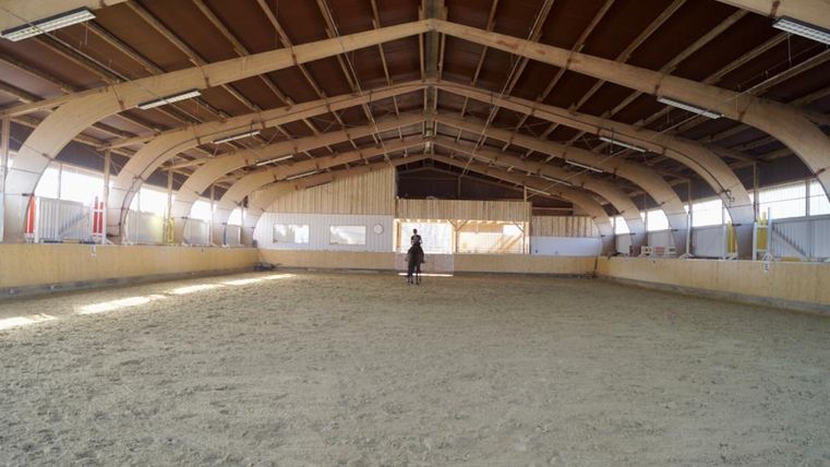 Une grande salle d'équitation avec un sol sablonneux. À l'arrière-plan, on voit un cavalier qui s'entraîne sur un cheval.